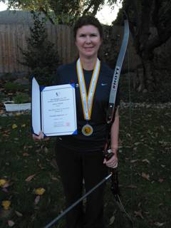 Janice with silver medal around her neck.  She is wearing a navy jacket with the US archery logo. She is holding her 3rd place qualifying award, and her bow is leaning at her side.