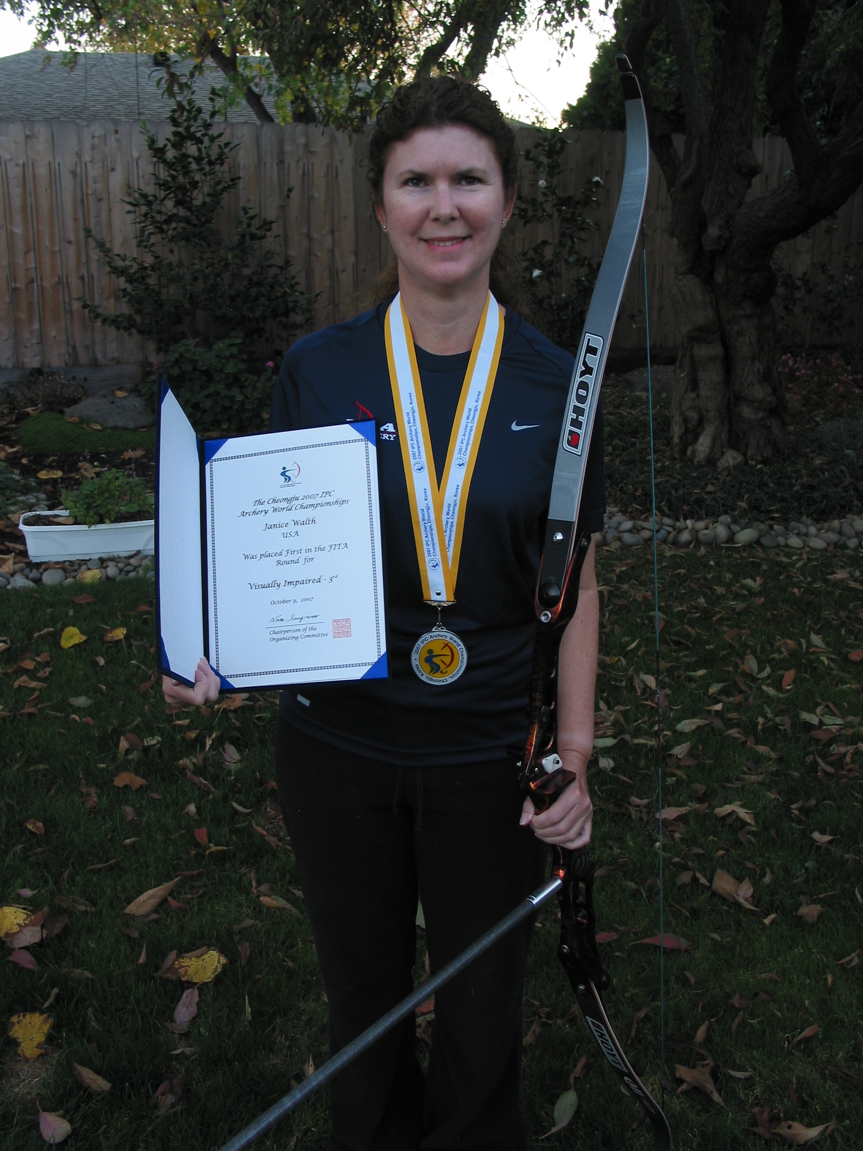 Janice Standing with silver medal around her neck. Her Hoyt bow is in her left hand and her award for the qualifier round is in her right hand
