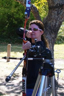 Photo of Janice at full draw.  She is lining up her shot by touching the back of her hand to the sight mounted on the tripod. 